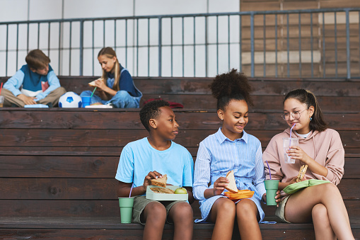 Group of three happy multicultural school friends having sandwiches and soda for lunch while sitting in row on wooden staircase