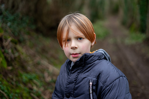 A young boy looks seriously into the camera. A boy of nine years old with long red hair in a warm outdoors jacket.