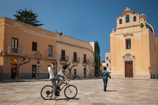 Horizontal shot of two young men riding a bike in Madrice Square with Madre Maria SS. Immacolata Church in the background, Flavignana, Sicily