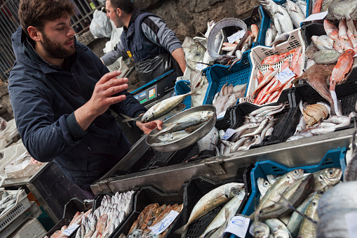 Horizontal shot of a fish vendor putting some fishes in the weighing scales in La Pescheria, the popular fish market, Catania, Sicily