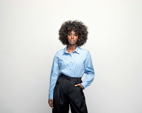 Young woman wearing blue shirt and black pants standing with hand in pocket and looking at camera. Studio shot on grey background.