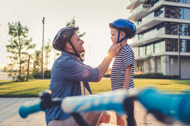 Father helping cheerful son wearing helmet for cycle. Excited little boy getting ready by wearing bike helmet to start cycling. Happy cute boy learn to ride a bike with his dad. Father helping cheerful son wearing helmet for cycle. Excited little boy getting ready by wearing bike helmet to start cycling. Happy cute boy learn to ride a bike with his dad. cycling helmet stock pictures, royalty-free photos & images