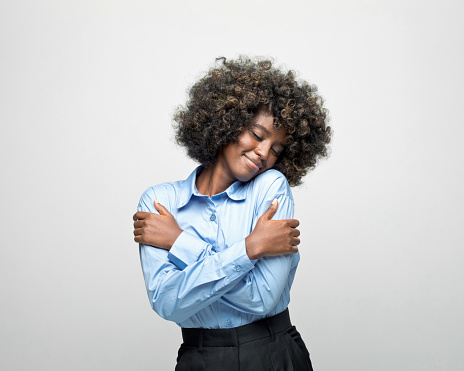 Young woman wearing blue shirt embracing herself and smiling. Studio shot on grey background.