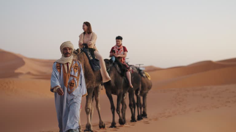 Asian Chinese Female Tourist Camel caravan going through the Sahara desert in Morocco at sunset