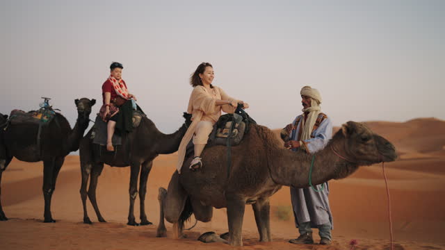 Asian Chinese female tourists getting up riding dromedary camel train crossing Sahara Desert Morocco