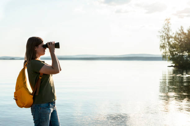 Young woman with yellow backpack looking through binoculars at birds on lake or river Birdwatching, zoology, ecology Research in nature, observation of animals Ornithology wildlife manager copy space mujer joven con mochila amarilla mirando a través de binoculares a pájaros en el lago o río observación de aves, zoología, ecología investigación en la naturaleza, observación de animales ornitología administrador de vida silvestre copiar espacio