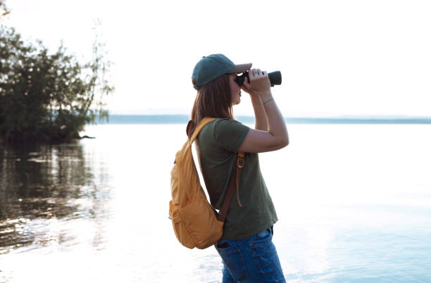 istock Young woman with yellow backpack looking through binoculars at birds on lake or river Birdwatching, zoology, ecology Research in nature, observation of animals Ornithology wildlife manager 1468218408