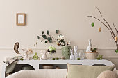 Interior design of easter living room interior with mock up poster frame, stylish sideboard, colorful easter eggs, easter bunny sculpture, vase with leaves and personal accessories Home decor Template