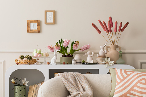 Interior design of easter living room interior with mock up poster frame, stylish white sideboard, hyacinth, easter bunny, vase with dried flowers and personal accessories. Home decor. Template.