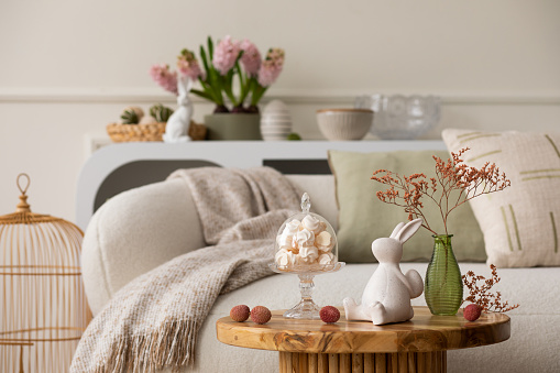Warm and cozy composition of easter living room interior with coffee table, white sofa, colorful easter eggs, easter bunny, hyacinth, vase with branch and personal accessories. Home decor. Template.
