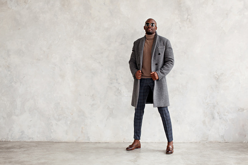 Handsome fashion man in turtleneck, gray coat, plaid trousers and leather loafer boots stands isolated on wall background, copy space. Fashionable male model