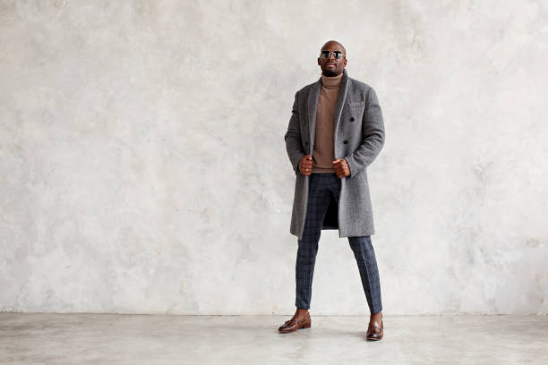 Handsome fashion man in turtleneck, gray coat, plaid trousers and leather loafer boots stands isolated on wall background, copy space. Fashionable male model stock photo