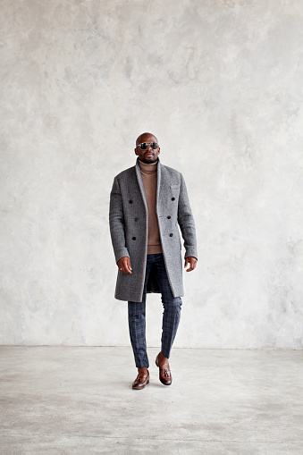 Full length of handsome young man in turtleneck, gray coat, plaid trousers and leather loafer boots walking isolated on wall background, copy space. Fashion style for men