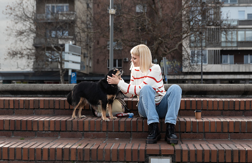 Young beautiful hipster woman is sitting on the stairs in the street with her foster dog she adopted from an animal shelter giving him love and attention