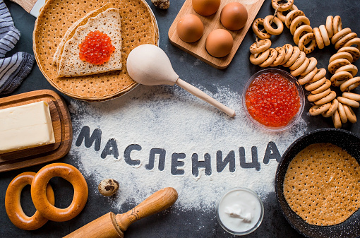 Russian traditions. Russian holiday Maslenitsa. A stack of pancakes, red caviar, bagels, bagels, a wooden spoon, eggs and butter. The Russian phrase Maslenitsa is written with flour. View from above.