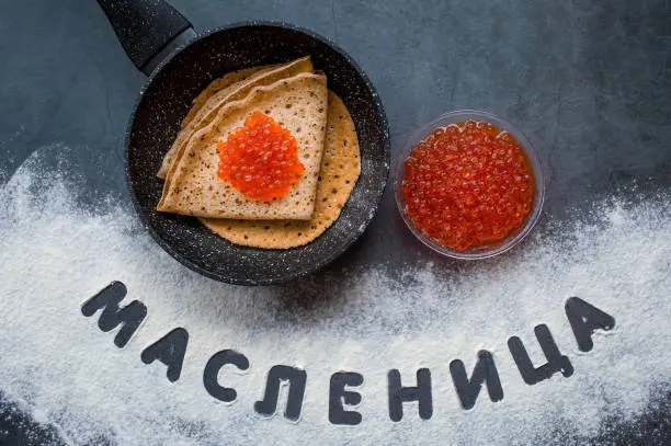 Russian traditions. Broad Maslenitsa. Smile. Frying pan, pancakes and red caviar. The inscription in Russian is "Maslenitsa". Written in flour. View from above.