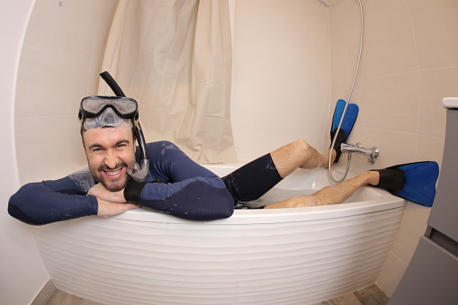 A humorous man in his forties is snorkeling in the bathtub. He has a beard and is home during the vacations