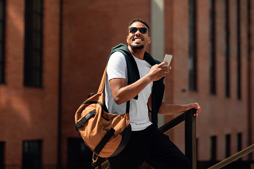 Happy trendy black guy in sunglasses using cellphone while walking down the street in urban city, smiling and looking away. Stylish man spending time outdoors