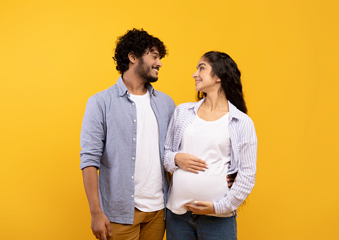 Happy indian couple expecting baby, posing over yellow background, embracing and looking at each other. Positive young pregnant family hugging and smiling