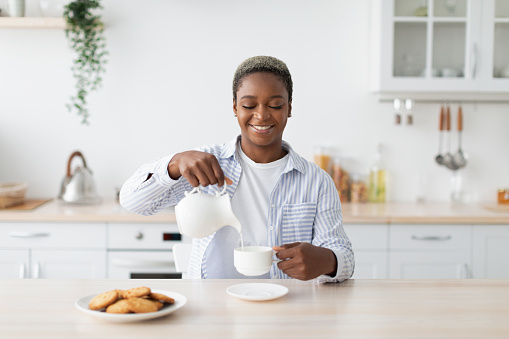Smiling millennial african american woman in casual pouring milk at coffee at table minimalist kitchen interior. Food blog, housewife rest at home, visit and healthy breakfast during covid-19 outbreak