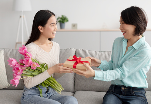 Happy mature Asian woman giving her adult daughter flowers and gift box, congratulating her with Woman's Day at home. Happy multi generation family enjoying holiday together in living room