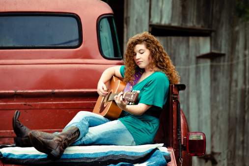 Young Woman plays guitar in the bed of a classic Chevy Pickup.  She is looking at the fret while she picks