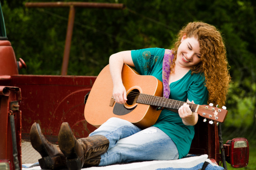 Young Woman plays guitar in the bed of a classic Chevy Pickup. She is smiling while her hands pick a tune