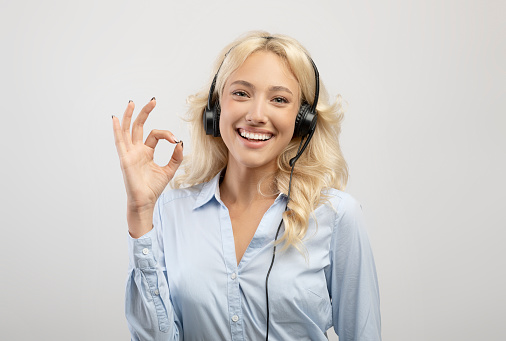 Happy female call centre operator with headset showing okay gesture and smiling at camera on light studio background. Positive technical support specialist looking at camera and expressing approval