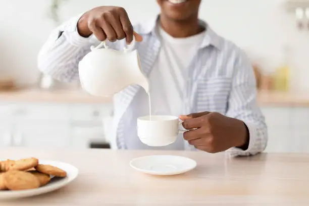 Photo of Satisfied young black woman pours milk at cup of favorite drink, sits at table with cookies in kitchen interior