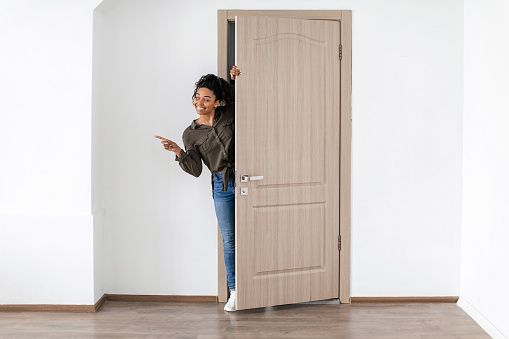 Cheerful African American Female Pointing Finger Looking Aside Opening Door Entering New House, Full Length Shot. Great Real Estate Offer. Look There Concept