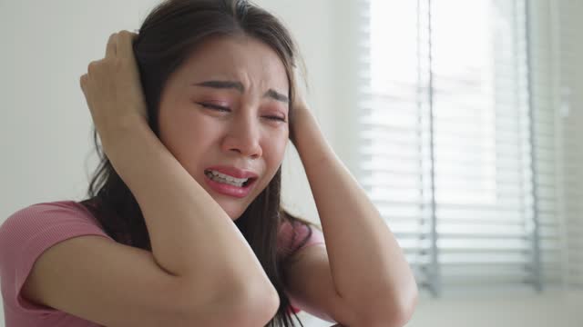 Asian beautiful upset depressed girl sitting alone on bed in bedroom. Attractive unhappy young woman feeling very sad, lonely and upset with life problem then crying with tears in quiet room at house.