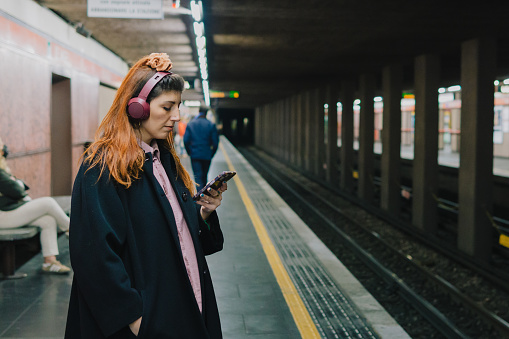 Bored young woman waits for subway listening music in headphones from smart phone