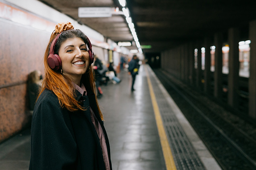 Young woman in the subway waits listening to music and looks into the camera smiling