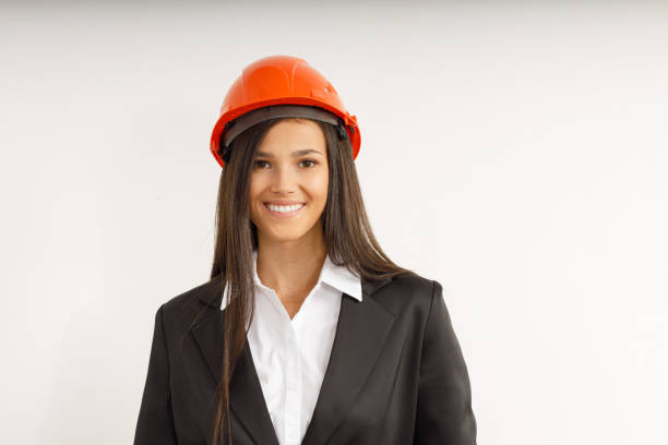 Beautiful model in orange construction helmet. Portrait of happy smiling brunette girl with long hair, dressed in strict suit. Studio shot of young business woman on isolated white background.
