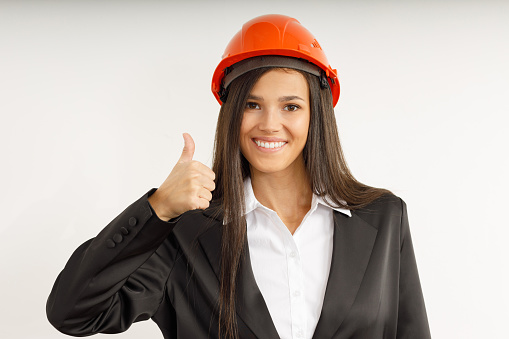 Beautiful model in orange construction helmet. Portrait of happy smiling brunette girl with thumbs up, dressed in strict suit. Studio shot of young business woman on isolated white background.