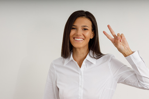 Happy young brunette woman in white shirt on isolated background. Portrait of attractive smiling girl looking straight into the camera and showing peace or victory sign with her fingers. Two fingers.