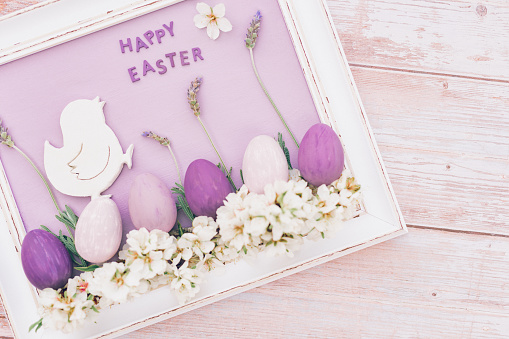 Flat lay of a white vintage picture frame with a pastel purple surface and a white happy easter chick with four lavender stems, almond blossoms and pink and purple colored easter eggs and with the English text happy easter in it. Vintage wooden background. Copy space on the rigth side. Color editing with added grain. Very selective and soft focus. Part of a series.