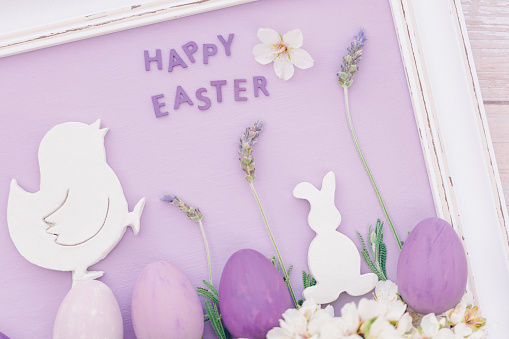 Flat lay of a part of a still life of a white vintage picture frame with a pastel purple surface and a white happy easter chick and easter bunny with four lavender stems, almond blossoms and pink and purple colored easter eggs and with the English text happy easter in it. Color editing with added grain. Very selective and soft focus. Part of a series.