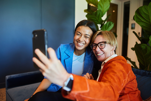 Two diverse businesswomen smiling during a video meeting on a smart phone while sitting on a sofa in an office lounge