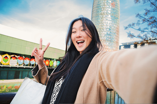 Happy asian young lady taking a selfie photo with an smartphone. Self portrait of chinese smiling woman doing the peace sign after shopping outdoors. High quality photo