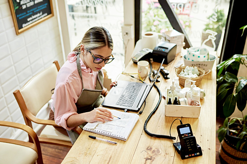 Woman wearing eyeglasses and apron over casual clothing, sitting at desk doing bookkeeping.