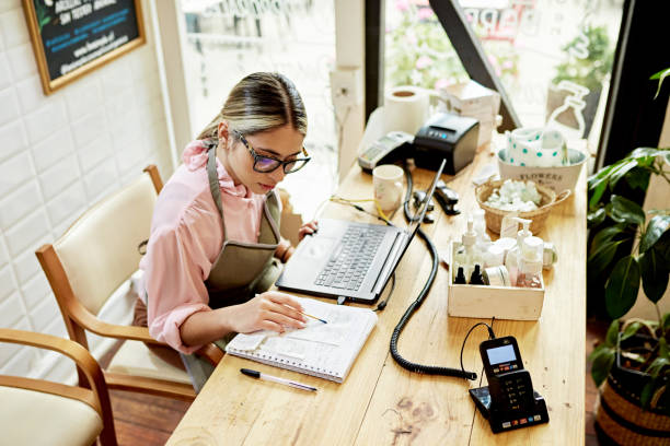 Young business owner using laptop in retail shop Woman wearing eyeglasses and apron over casual clothing, sitting at desk doing bookkeeping. small business stock pictures, royalty-free photos & images