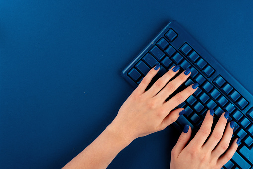 Woman hands typing on computer keyboard on classic blue background, top view