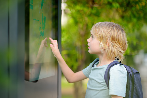 Little boy tourist studying at the big public outdoor street map of city. Kid touching screen and looking for the way. Child is interested in a visual map or advertising billboards on the urban street