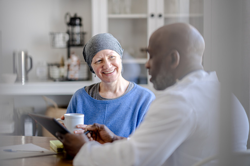 A male Oncologist of African decent sits with a senior woman in the comfort of her own home during a Homecare visit.  The patient is dressed comfortably and wearing a headscarf to keep her warm as the two talk about her Cancer treatment.