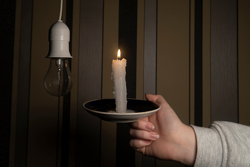 A hand with a burning candle next to a cartridge with an electric light bulb in a dark room (close-up). Blackout. Power outage concept