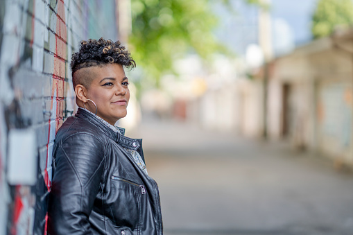 A young woman of African American decent, poses for a portrait while enjoying the outdoors.  She is dressed in a trendy leather jacket and smiling as she explores the downtown core.