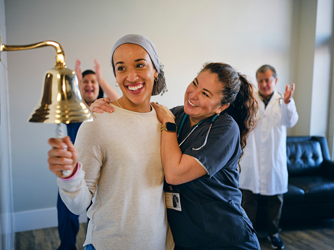 An adult woman chemotherapy patient in a treatment office, celebrating the completion of her treatment with a ceremonial bell ring. Actor portrayal.