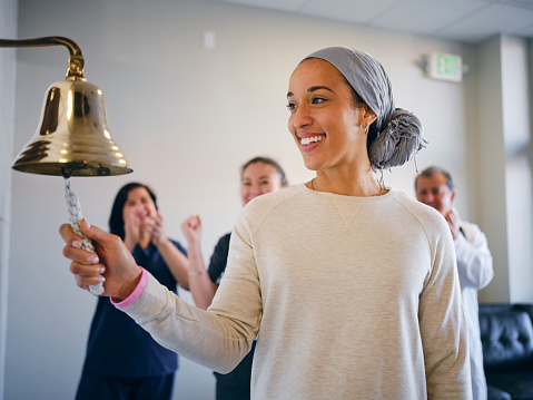 An adult woman chemotherapy patient in a treatment office, celebrating the completion of her treatment with a ceremonial bell ring. Actor portrayal.