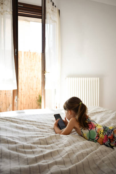 vue arrière d’une petite fille allongée sur un lit et utilisant un téléphone portable. - telephone window mobile phone addiction photos et images de collection